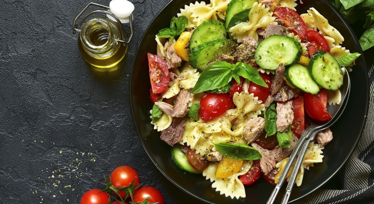Ein Teller mit Nudelsalat aus Farfalle, Tomaten, Gurken und Thunfisch auf dunklem Untergrund, daneben eine Flasche Olivenöl und Kirschtomaten.