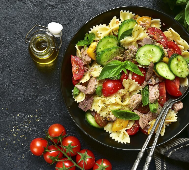 Ein Teller mit Nudelsalat aus Farfalle, Tomaten, Gurken und Thunfisch auf dunklem Untergrund, daneben eine Flasche Olivenöl und Kirschtomaten. | © Getty Images/Lilechka75