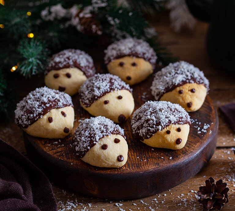 Mehrere mit Kokosraspel dekorierte Igelkekse auf einem Holzbrett in weihnachtlichem Ambiente.  | © Shutterstock/ A. Zhuravleva