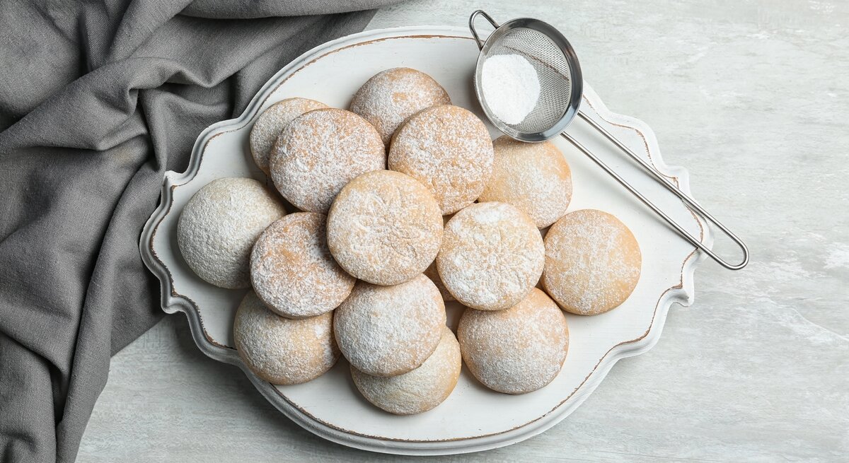Mehrere mit Puderzucker bestäubte Butterwölkchen-Plätzchen auf weißem Servierteller.