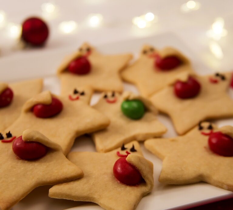 Niedliche "Himmelsboten"-Plätzchen mit aufgemalten Gesichtern, die farbige Schokolinsen „umarmen“, auf weißem Teller arrangiert. | © Shutterstock/S C.Welman