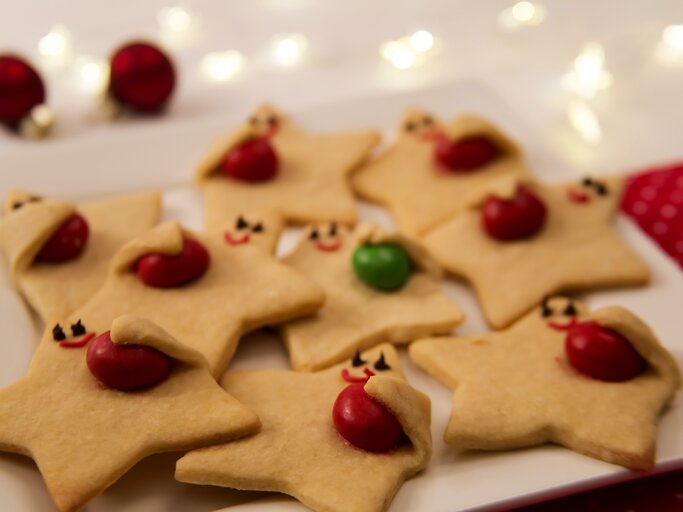 Niedliche "Himmelsboten"-Plätzchen mit aufgemalten Gesichtern, die farbige Schokolinsen „umarmen“, auf weißem Teller arrangiert. | © Shutterstock/S C.Welman