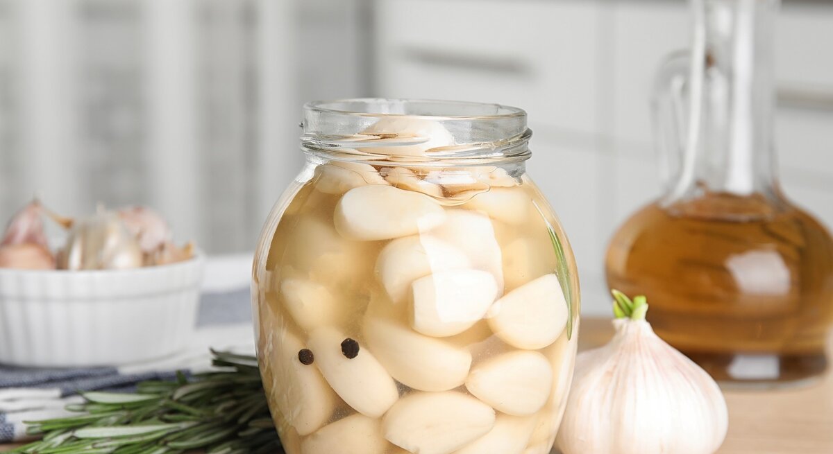 In einem hellen Küchenumfeld stehen ein Glas mit eingelegten Knoblauchzehen und eine Knoblauchknolle neben Rosmarin und Ölflasche.