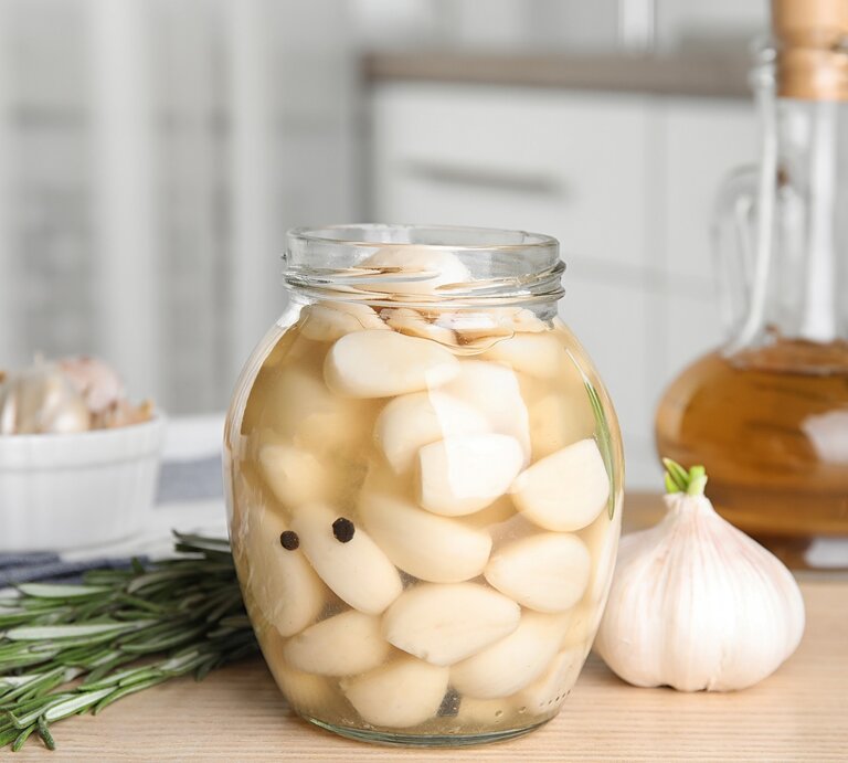In einem hellen Küchenumfeld stehen ein Glas mit eingelegten Knoblauchzehen und eine Knoblauchknolle neben Rosmarin und Ölflasche. | © Shutterstock/New Africa