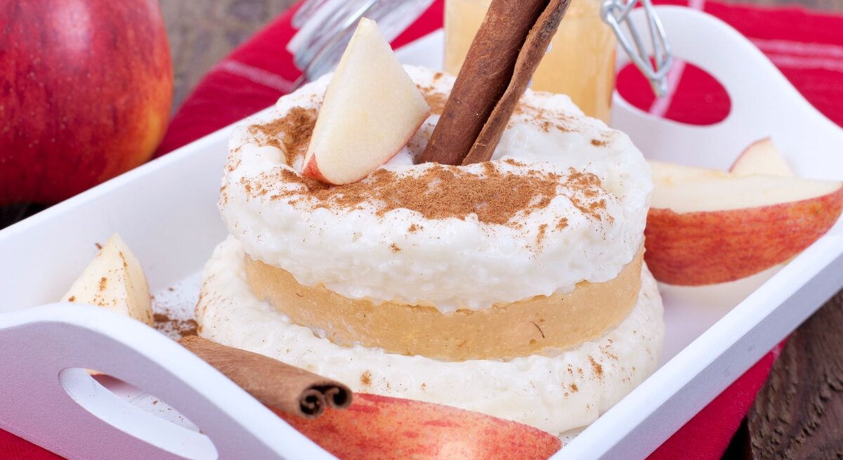 Ein cremiger Milchreis mit Apfelmus, Zimtstange und Apfelspalten auf weißem Tablett, auf rotem Tuch und Holztisch.