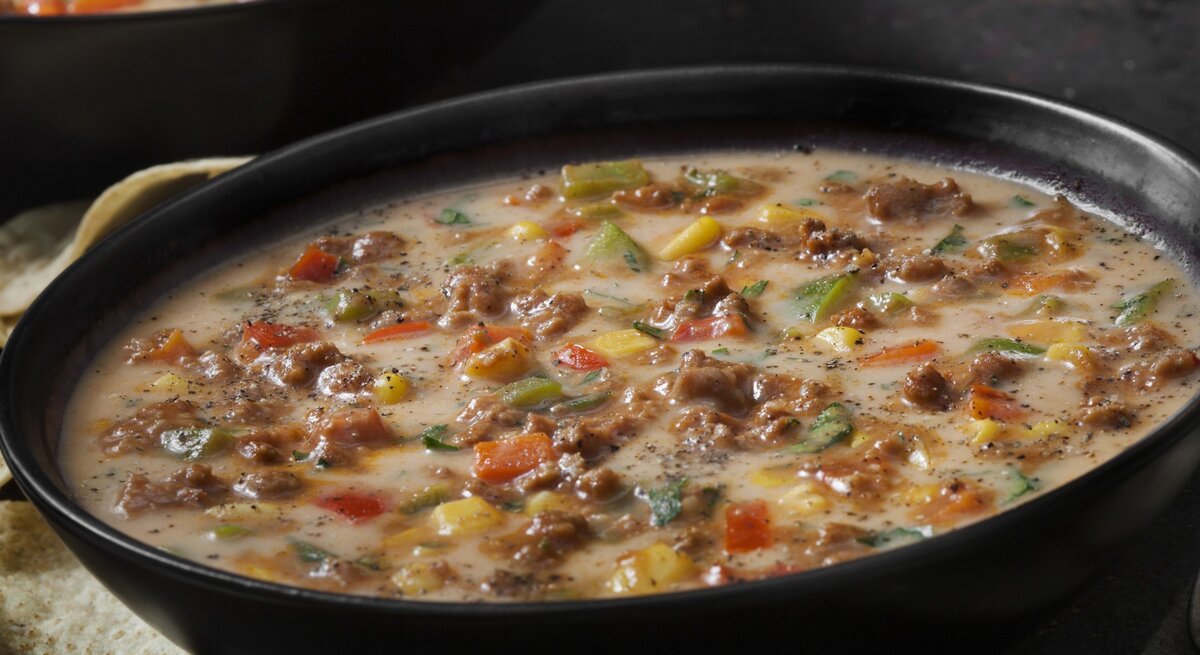 Das Bild zeigt eine herzhafte Schüssel Suppe, die mit Hackfleisch, Käse und verschiedenen Gemüsesorten wie Paprika, Zwiebeln und Mais zubereitet wurde. Die Suppe scheint cremig zu sein und ist großzügig mit schwarzen Pfeffer bestreut, was auf eine würzige Note hindeutet. 