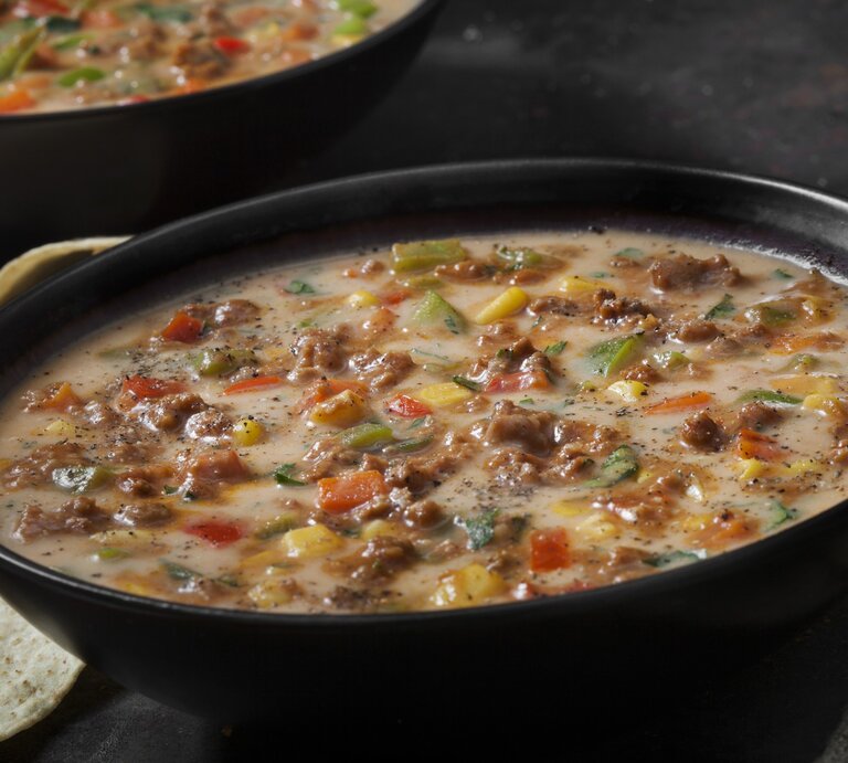 Das Bild zeigt eine herzhafte Schüssel Suppe, die mit Hackfleisch, Käse und verschiedenen Gemüsesorten wie Paprika, Zwiebeln und Mais zubereitet wurde. Die Suppe scheint cremig zu sein und ist großzügig mit schwarzen Pfeffer bestreut, was auf eine würzige Note hindeutet.  | © GettyImages/LauriPatterson