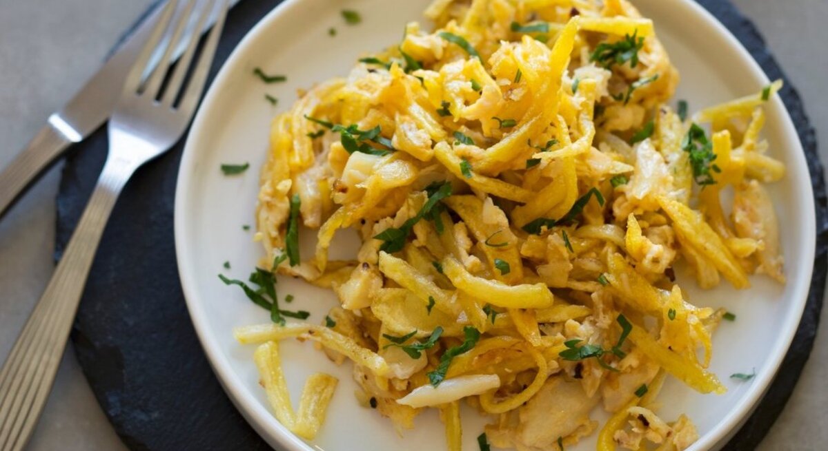 Alt-Text: Ein Teller mit Bacalhau à Brás, einem traditionellen portugiesischen Gericht aus zerpflücktem Stockfisch, der mit dünn geschnittenen, goldgelben Kartoffelstreifen und Eiern vermischt ist. Es ist garniert mit frischer, grüner Petersilie, die einen schönen Farbkontrast bildet. 