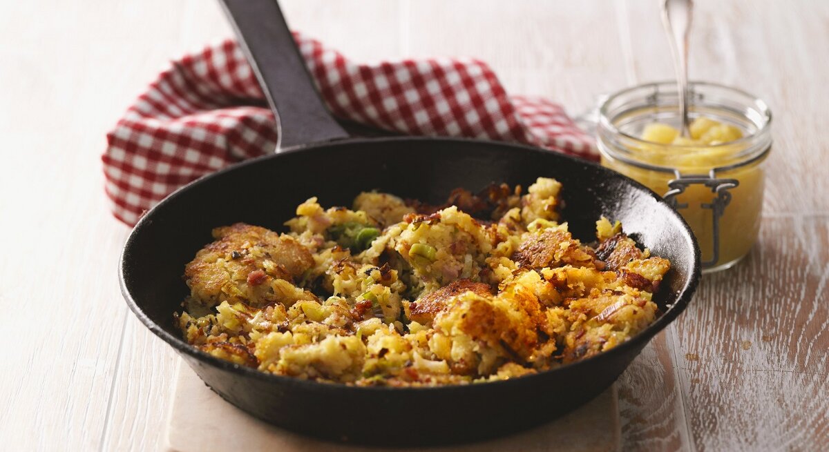 Eine Pfanne mit einem Pfannengericht mit Kartoffeln steht auf einer Holztischplatte. Daneben ein kleines Glas mit Apfelmus.