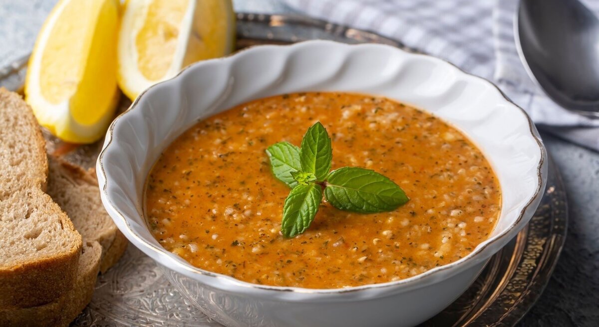 Ein Bild zeigt eine Schüssel mit Tarhana-Suppe, einer traditionellen türkischen Suppe, die reich an Gewürzen und Tomaten ist. Die Suppe hat eine leuchtend orangefarbene Textur mit sichtbaren Kräutern und Körnern, die eine rustikale und nahrhafte Mahlzeit suggerieren. Die Schüssel steht auf einem dekorativen Metalltablett und wird von zwei Zitronenspalten, einem Stapel Vollkornbrot und einem Löffel, der auf einem Stoffserviette liegt, flankiert. Ein Zweig frische Minze ziert die Mitte der Suppe, was einen farblichen Kontrast und einen Hinweis auf Frische gibt.