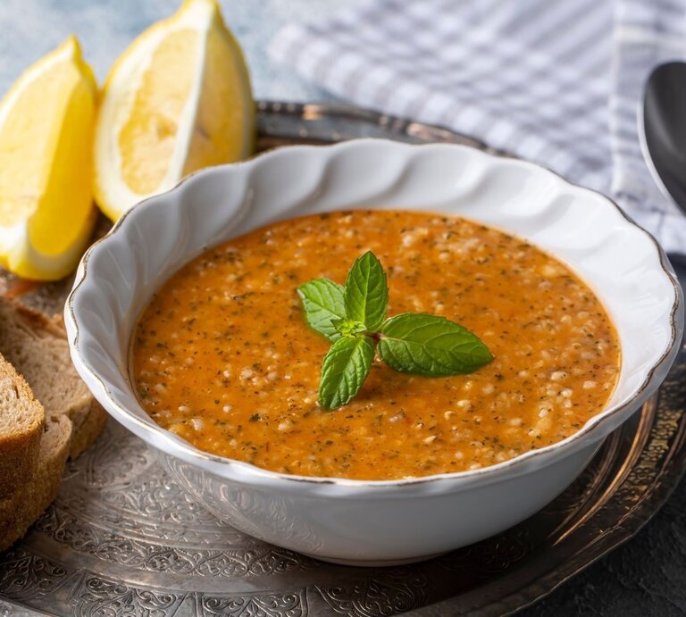 Ein Bild zeigt eine Schüssel mit Tarhana-Suppe, einer traditionellen türkischen Suppe, die reich an Gewürzen und Tomaten ist. Die Suppe hat eine leuchtend orangefarbene Textur mit sichtbaren Kräutern und Körnern, die eine rustikale und nahrhafte Mahlzeit suggerieren. Die Schüssel steht auf einem dekorativen Metalltablett und wird von zwei Zitronenspalten, einem Stapel Vollkornbrot und einem Löffel, der auf einem Stoffserviette liegt, flankiert. Ein Zweig frische Minze ziert die Mitte der Suppe, was einen farblichen Kontrast und einen Hinweis auf Frische gibt. | © Shutterstock/Esin Deniz