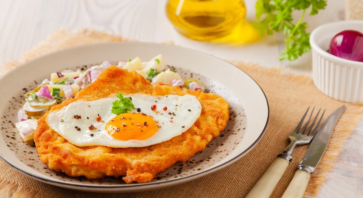 Ein appetitliches Wiener Schnitzel, gekrönt von einem Spiegelei mit einem glänzenden Dotter, liegt auf einem Teller mit fein gewürfelten Zwiebeln und Kräutern. Der Teller steht auf einer Stoffunterlage auf einem Holztisch. Im Hintergrund ist eine Flasche Olivenöl und ein Ramekin mit einer roten Zwiebel zu sehen. Ein Messer und eine Gabel mit Elfenbeingriffen liegen neben dem Teller