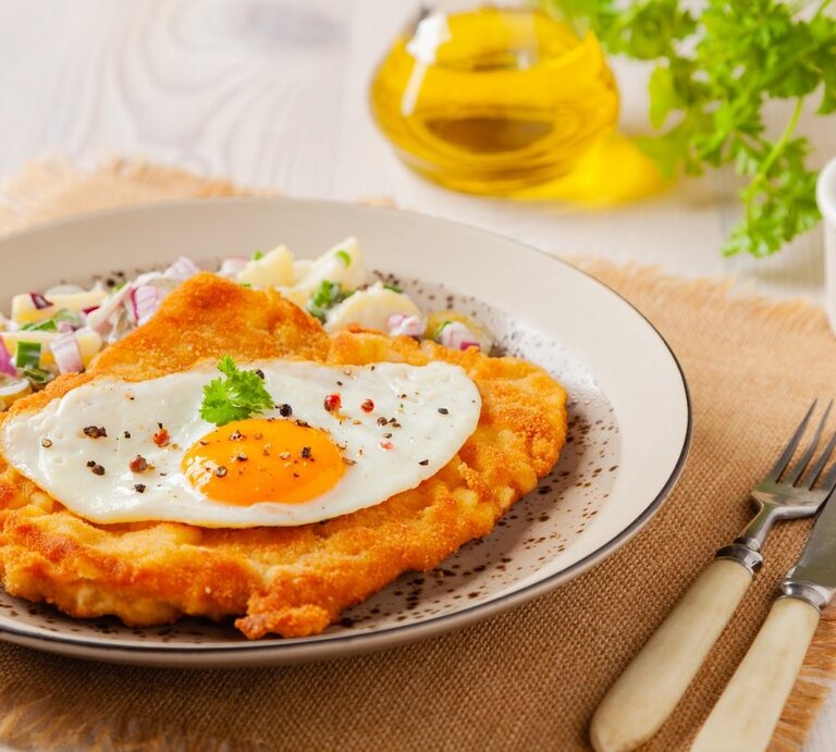 Ein appetitliches Wiener Schnitzel, gekrönt von einem Spiegelei mit einem glänzenden Dotter, liegt auf einem Teller mit fein gewürfelten Zwiebeln und Kräutern. Der Teller steht auf einer Stoffunterlage auf einem Holztisch. Im Hintergrund ist eine Flasche Olivenöl und ein Ramekin mit einer roten Zwiebel zu sehen. Ein Messer und eine Gabel mit Elfenbeingriffen liegen neben dem Teller | © AdobeStock/gkrphoto