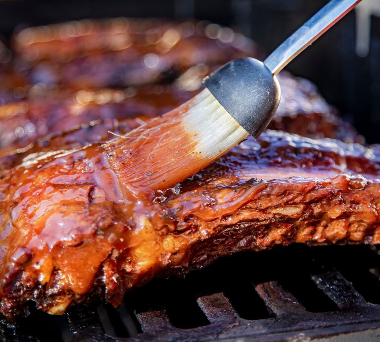 Die Spareribs von der Seite auf dem Grill fotografiert, während sie gerade mit Marinad eeingepinselt werden. | © Shutterstock/ F Armstrong Photography