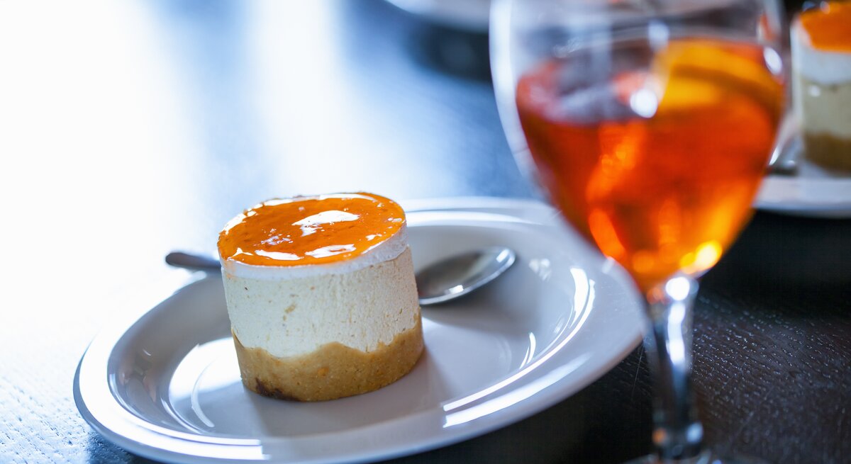 Ein kleiner Aperol-Käsekuchen mit einem orangenen Guss aus Orangensaft, Prosecco und Aperol neben einem Glas mit Aperol Spitz