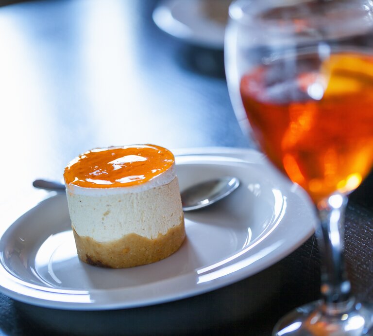 Ein kleiner Aperol-Käsekuchen mit einem orangenen Guss aus Orangensaft, Prosecco und Aperol neben einem Glas mit Aperol Spitz | © Shutterstock/ NatashaPhoto