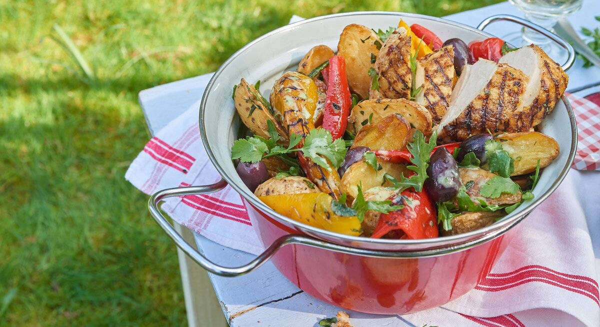 Eine rote Schüssel mit dem Grillkartoffelsalat mit Hähnchen. Alles auf einem Tisch mit weißer Tischdecke und rötlicher Deko