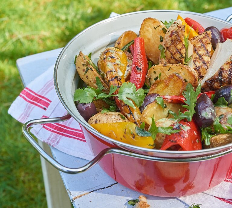 Eine rote Schüssel mit dem Grillkartoffelsalat mit Hähnchen. Alles auf einem Tisch mit weißer Tischdecke und rötlicher Deko | © Matthias Liebich
