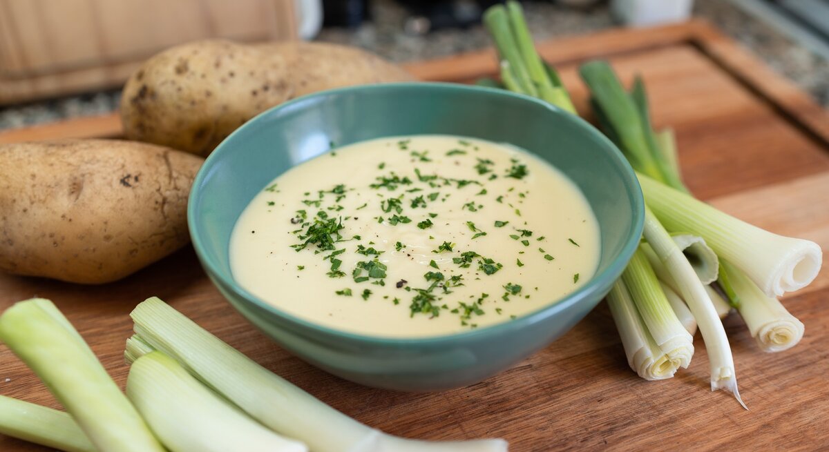 Eine blaue Suppenschale mit der samtigen Suppe auf einem Holztisch. Drum herum Kartoffeln und Lauch.