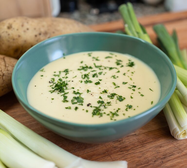Eine blaue Suppenschale mit der samtigen Suppe auf einem Holztisch. Drum herum Kartoffeln und Lauch. | © Shutterstock/ KGBR