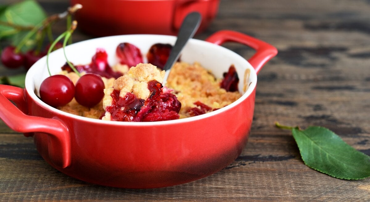 Eine kleine rote Backform mit dem Kirsch-Crumble auf einem Holztisch. | © GettyImages/ zefirchik06
