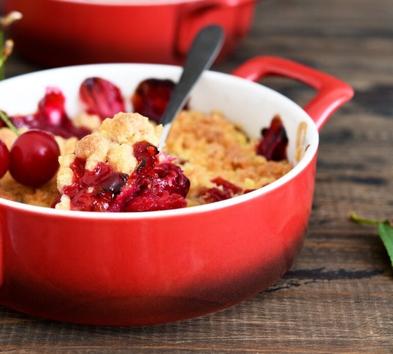 Eine kleine rote Backform mit dem Kirsch-Crumble auf einem Holztisch. | © GettyImages/ zefirchik06