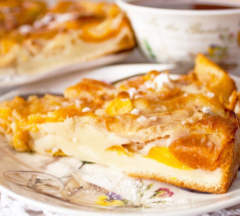 Ein Stück vom Pfirsichkuchen auf einem altmodischen Teller und hellem Holztisch. Alles von der Seite fotografiert. | © GettyImages/ Евгения Матвеец