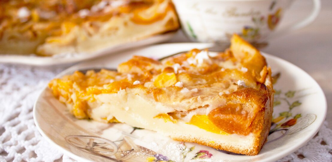 Ein Stück vom Pfirsichkuchen auf einem altmodischen Teller und hellem Holztisch. Alles von der Seite fotografiert. | © GettyImages/ Евгения Матвеец