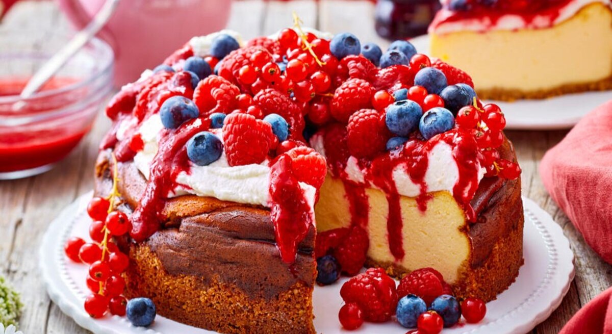 Zu sehen ist ein leckerer Käsekuchen mit Sahne-Beeren-Topping und präsentiert vor einem roten Hintergrund