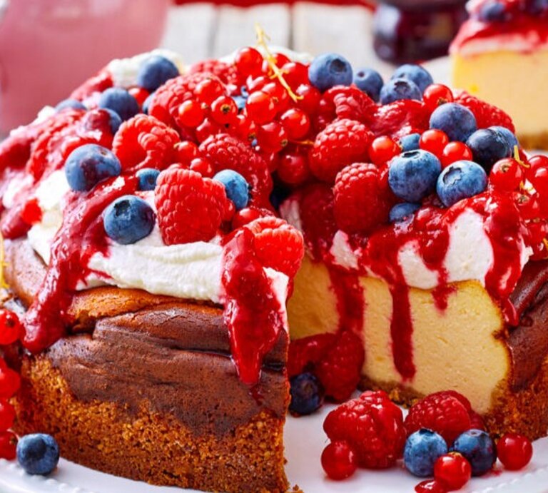 Zu sehen ist ein leckerer Käsekuchen mit Sahne-Beeren-Topping und präsentiert vor einem roten Hintergrund | © Eat Club