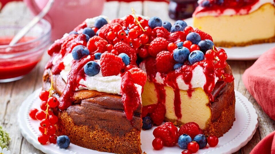 Zu sehen ist ein leckerer Käsekuchen mit Sahne-Beeren-Topping und präsentiert vor einem roten Hintergrund | © Eat Club
