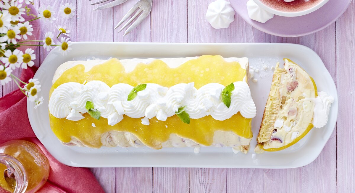 Der Eiskuchen auf einem violetten Tisch mit Blumendeko und auf länglichem weißen Teller angerichtet. Alles direkt von oben fotografiert.