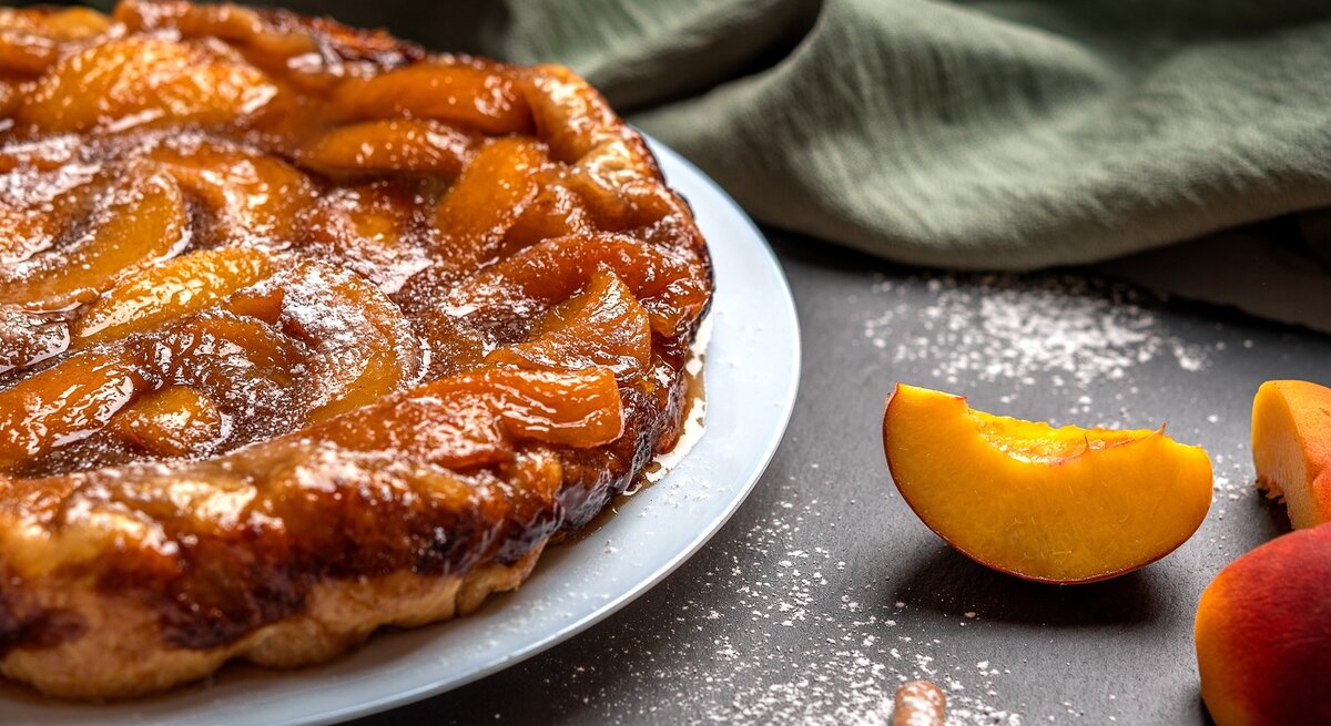 Die Tarte ist links im Bild sehr nah zu sehen und rechts daneben frische Pfirsichspalten. Alles auf dunklem Untergrund fotografiert.