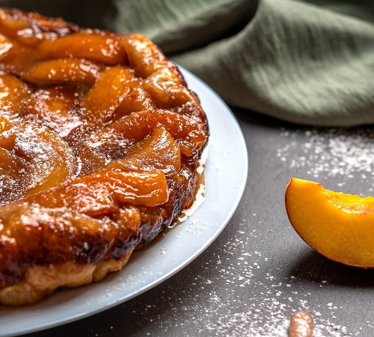 Die Tarte ist links im Bild sehr nah zu sehen und rechts daneben frische Pfirsichspalten. Alles auf dunklem Untergrund fotografiert. | © Shutterstock/ Vitaly Krivchikov