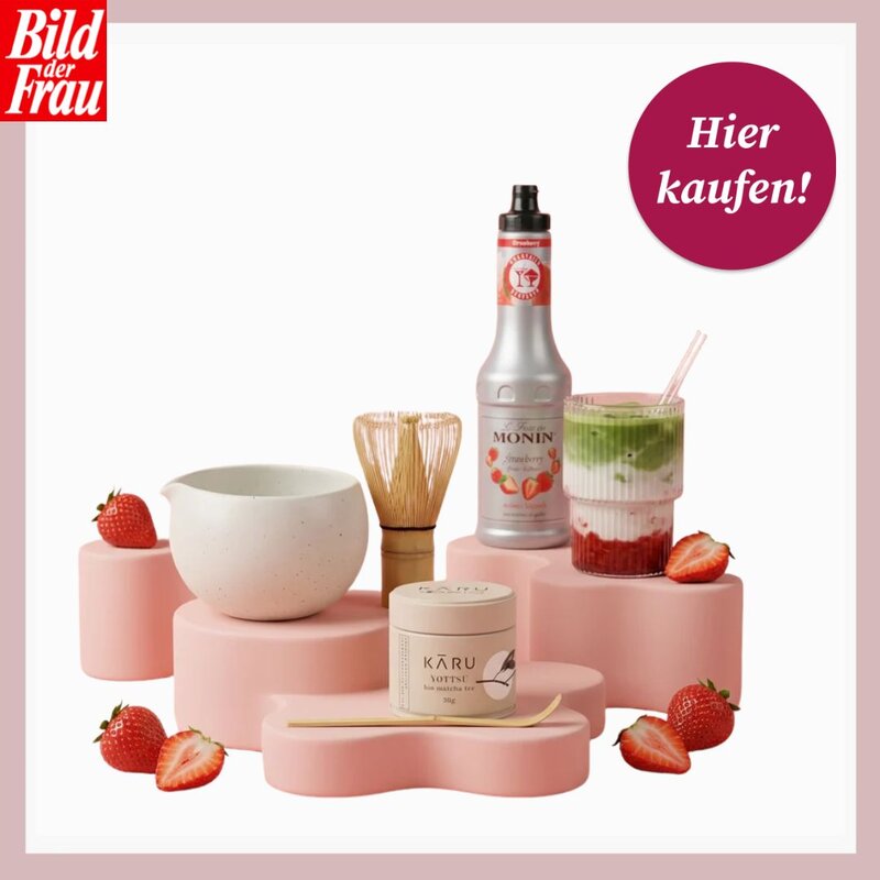Matcha-Latte mit Erdbeeren im Glas neben Sirupflasche, Matchabesen und Keramikschale auf rosafarbenen Podesten. | © Karu/Canva/Collage:bildderfrau.de