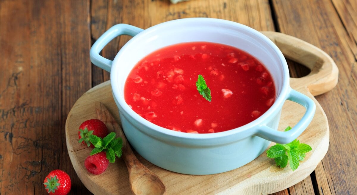 Eine hellblaue Suppenschüssel mit Henkeln gefüllt mit roter Erdbeerkaltschale, garniert mit einem Minzblatt, auf einem Holzbrett neben frischen Erdbeeren und Himbeeren.