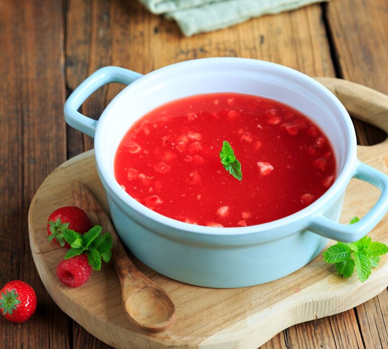 Eine hellblaue Suppenschüssel mit Henkeln gefüllt mit roter Erdbeerkaltschale, garniert mit einem Minzblatt, auf einem Holzbrett neben frischen Erdbeeren und Himbeeren. | © AdobeStock/Juefrateam