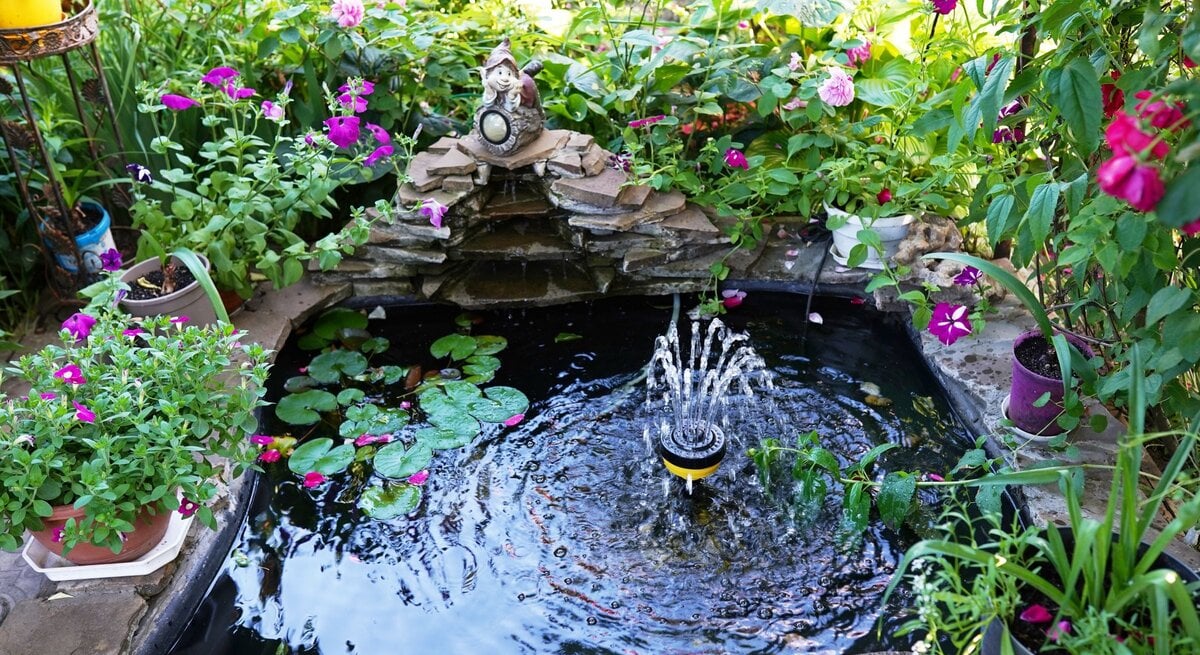 Üppig bepflanzter Garten mit kleinem Mini-Brunnen und sprudelnder Fontäne, umgeben von rosa Blüten, Seerosen und dekorativer Zwergenfigur.