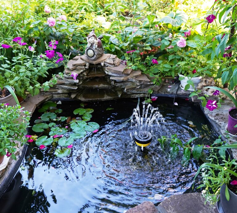 Üppig bepflanzter Garten mit kleinem Mini-Brunnen und sprudelnder Fontäne, umgeben von rosa Blüten, Seerosen und dekorativer Zwergenfigur. | © Shutterstock/nastya_ph