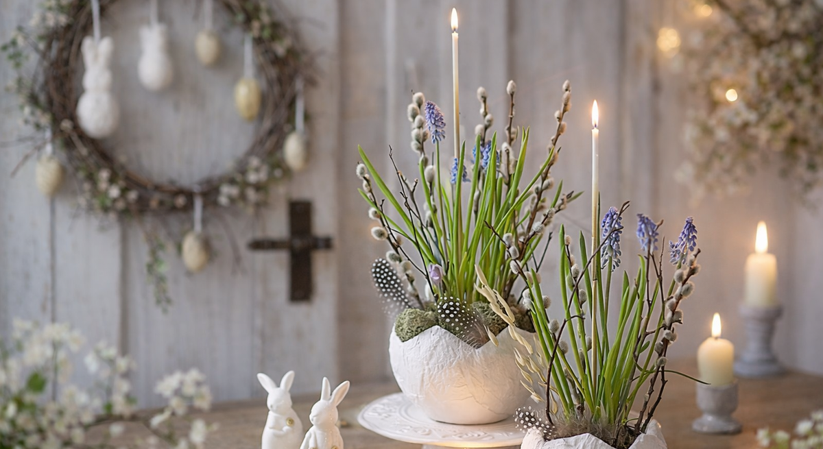 Frühlingshafte Osterdeko mit bepflanzter Eierschale aus Toilettenpapier, Kerzen, Weidenkätzchen und weißen Hasenfiguren auf Holztisch vor Kranz und Kreuz.
