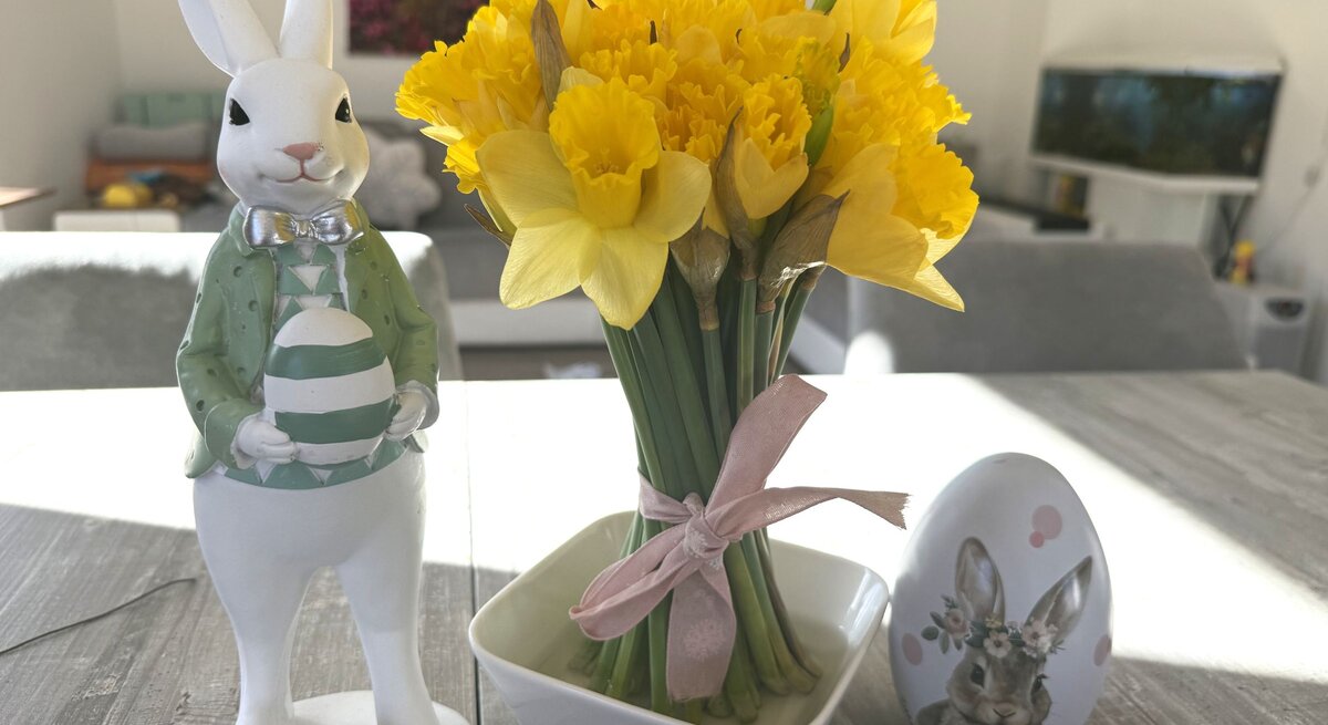Gelbe Narzissen in kleiner Vase mit rosa Schleife stehen auf Holztisch neben Osterhasenfigur mit Ei und bemaltem Deko-Ei im hellen Wohnzimmer.