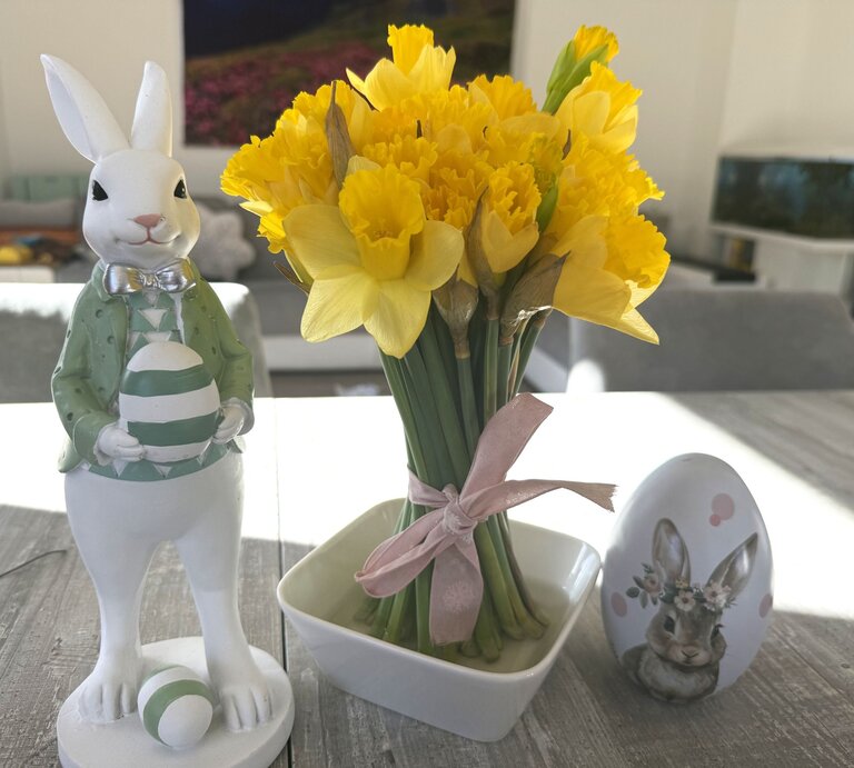 Gelbe Narzissen in kleiner Vase mit rosa Schleife stehen auf Holztisch neben Osterhasenfigur mit Ei und bemaltem Deko-Ei im hellen Wohnzimmer. | © Madeleine Liedke 