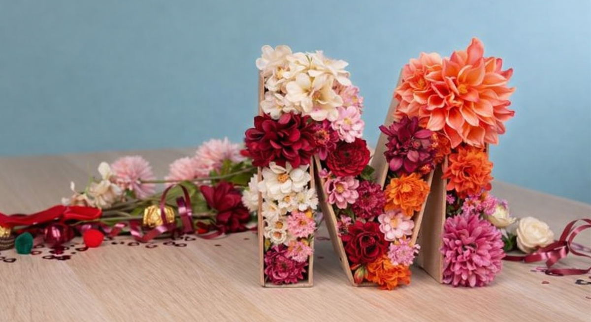Mit bunten Dahlien und Rosen gefüllter Buchstabe M aus Karton steht auf Holztisch vor hellblauem Hintergrund.