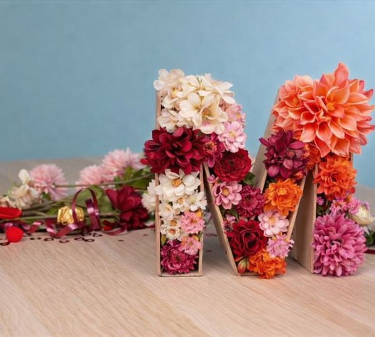 Mit bunten Dahlien und Rosen gefüllter Buchstabe M aus Karton steht auf Holztisch vor hellblauem Hintergrund. | © Generiert mit KI (Erstellt mit ChatGPT)