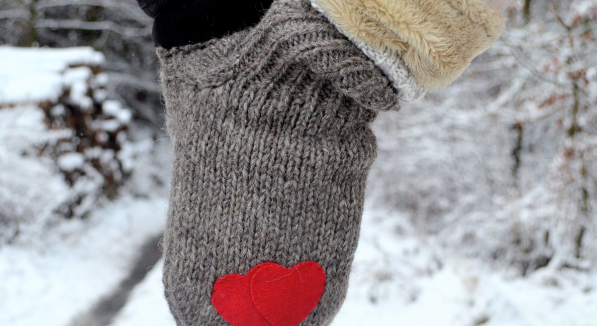 Zwei Hände in grauem Doppelhandschuh mit roten Herzen halten sich auf einem verschneiten Waldweg im Winter.