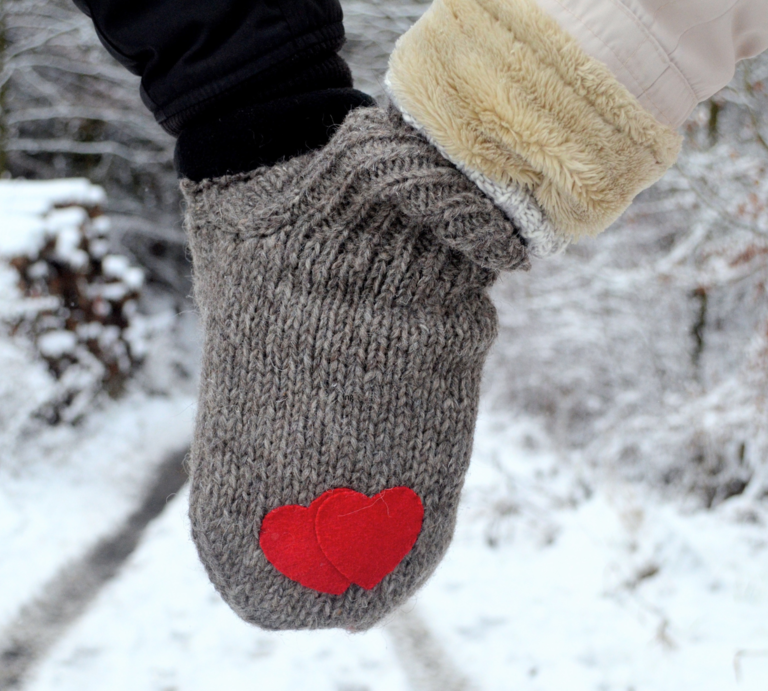 Zwei Hände in grauem Doppelhandschuh mit roten Herzen halten sich auf einem verschneiten Waldweg im Winter. | © Canva