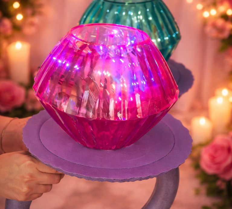 Ein selbst gebastelter Diamantring aus pinkem und türkisfarbenem Glas leuchtet auf einer lilafarbenen Halterung vor Kerzen und Rosen. | © Generiert mit KI (Erstellt mit ChatGPT)