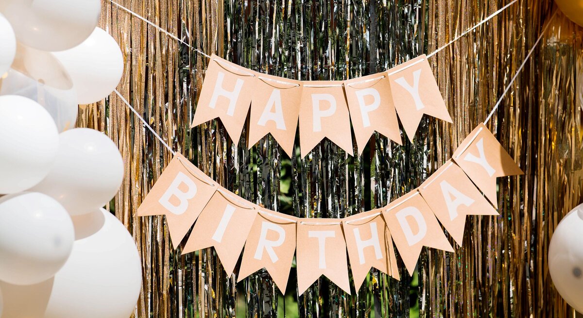 Geburtstagsgirlande basteln: Beige Wimpelkette mit weißem „Happy Birthday“-Schriftzug hängt festlich vor schimmerndem Goldvorhang mit Ballondekoration.