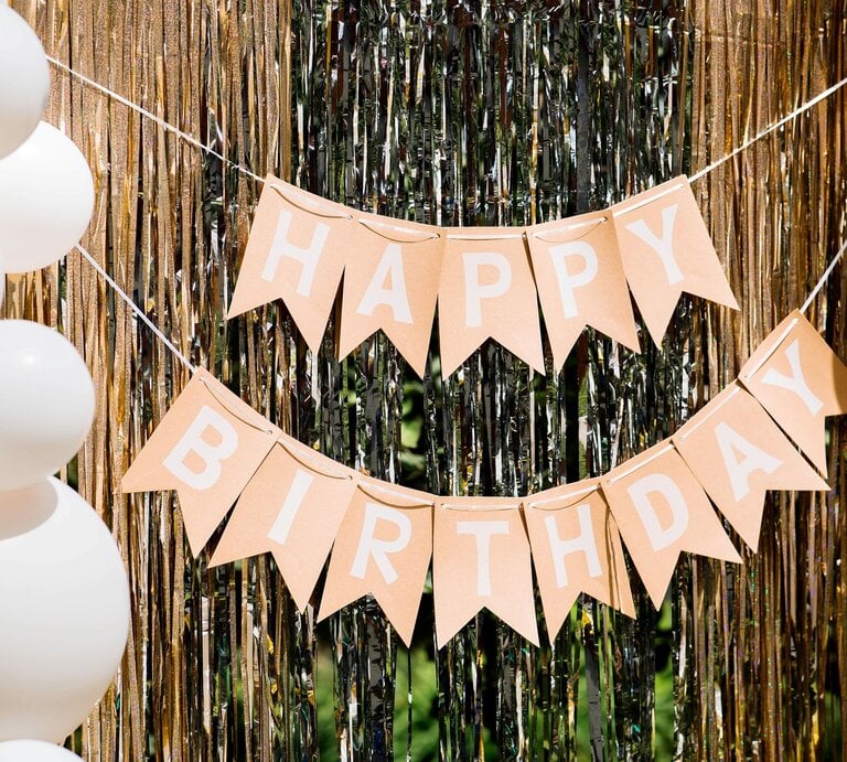 Geburtstagsgirlande basteln: Beige Wimpelkette mit weißem „Happy Birthday“-Schriftzug hängt festlich vor schimmerndem Goldvorhang mit Ballondekoration. | © Shuterstock/PhotoJuli86