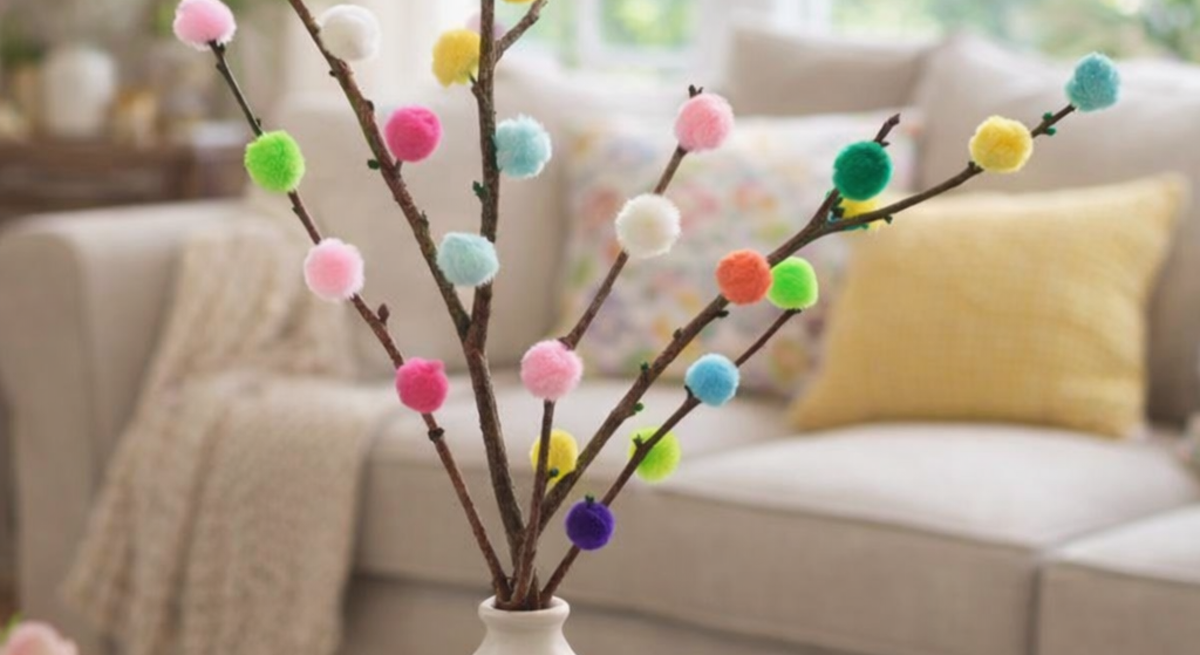 Farbige Pompons zieren mehrere Zweige in einer schlichten Vase auf einem Tisch, im Hintergrund ein gemütliches Sofa mit Kissen.